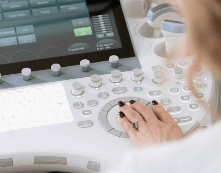 A woman operates a computer to manage an ultrasound machine, focused on the screen in a clinical setting.